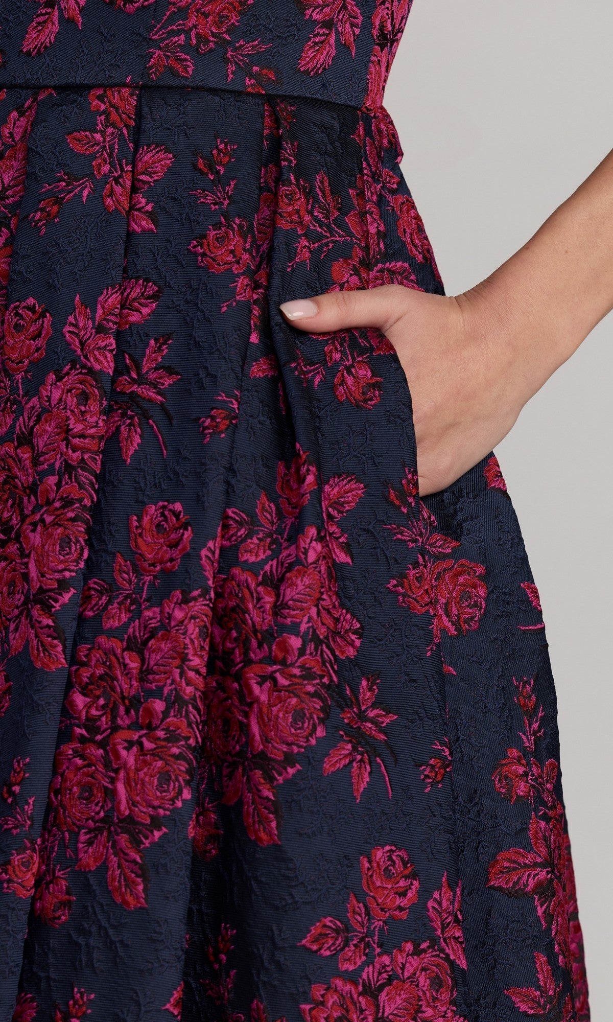A person with light nail polish places their hand in the pocket of the Mac Duggal 68625 tea-length party dress, a navy blue piece adorned with a vibrant pink floral pattern.